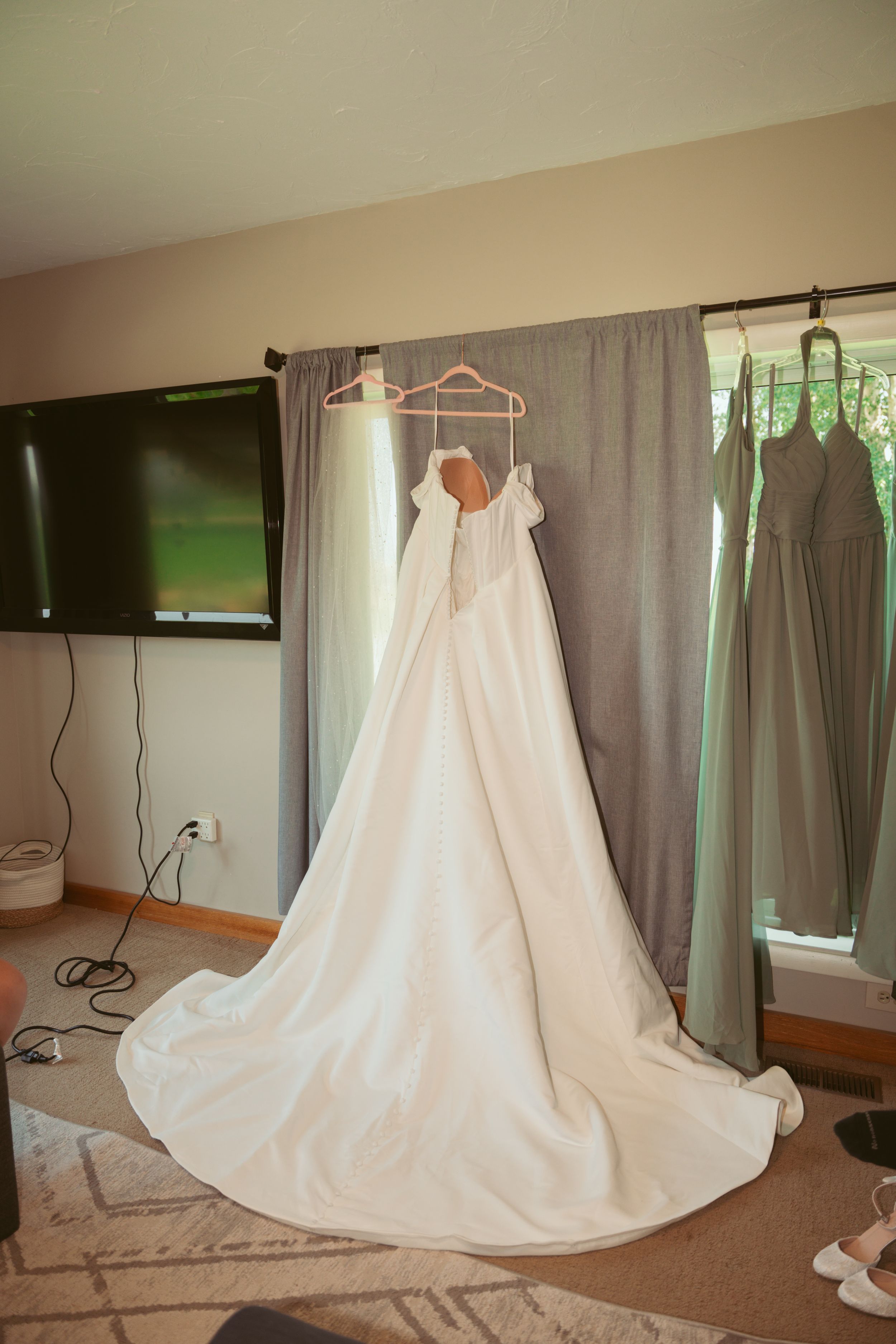 A white wedding dress with a long train hangs on display against grey curtains in a room with a mounted TV.