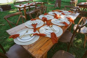 Rustic wooden tables set outdoors with white plates and orange bow decorations for an elegant outdoor event setting.