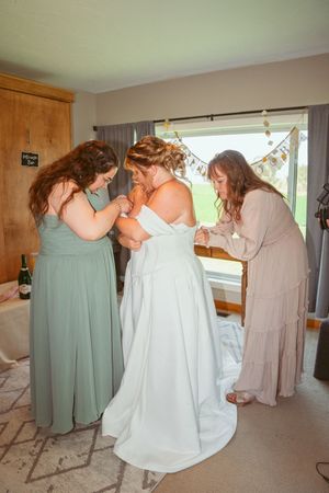 Bridesmaids in pastel dresses help with final wedding dress preparations in a sunlit room.