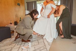 Wedding party members assist with adjusting the hem of a white wedding dress.