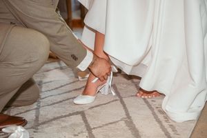 Close-up of white dress hemline and shoes on tiled floor during wedding preparations.