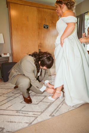 Someone assists with adjusting the bride's shoes as she stands in her elegant white wedding gown.