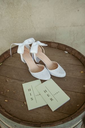 White bridal shoes with bow details and wedding cards displayed on a rustic wooden barrel.