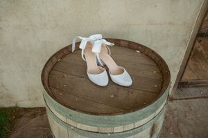 White bridal heels with bows placed on rustic wooden barrel outdoors.