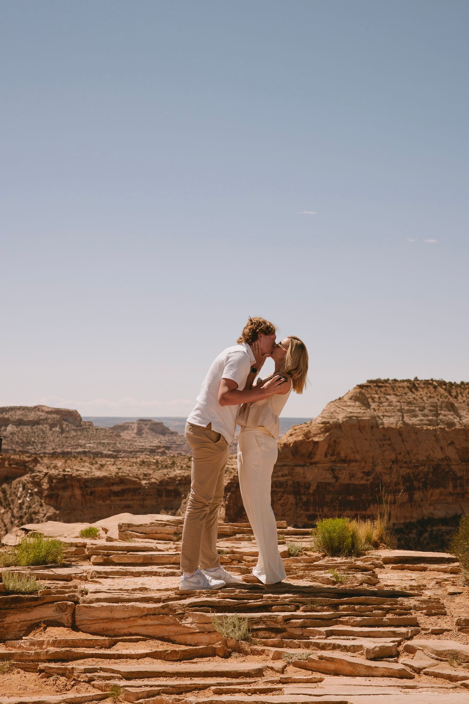 Two people embrace in white outfits while standing on red rock formations in a desert canyon landscape.
