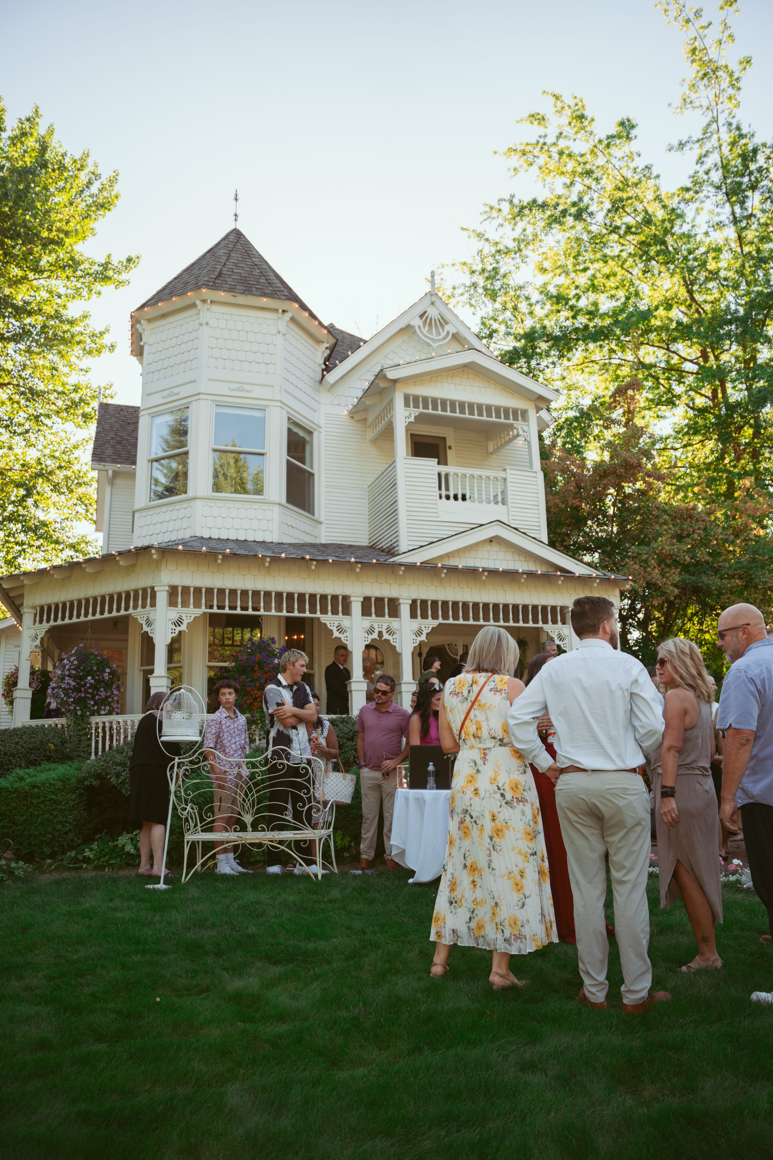 A group gathers on the lawn of a charming white Victorian home during a social event in the late afternoon.