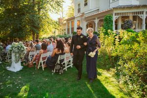 A garden wedding ceremony in progress with guests seated on white chairs as attendees walk down a lush outdoor aisle.