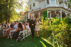 A beautiful outdoor wedding ceremony takes place in front of a Victorian-style house with white chairs on the lawn at sunset.
