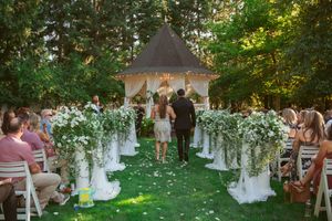 An outdoor wedding ceremony at a gazebo decorated with flowing white floral arrangements and guests seated on both sides.
