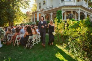 Outdoor wedding guests seated on white chairs on a lawn beside a Victorian-style house in warm evening sunlight.