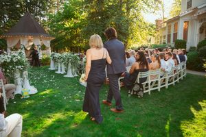 Guests gather on a sunlit lawn for an outdoor wedding ceremony with white chairs and natural greenery.