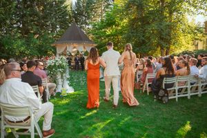 Wedding ceremony processional takes place on a sunny lawn with white chairs and floral arch at an outdoor garden venue.
