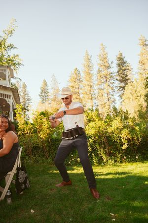 Person in cowboy hat and casual attire dances outdoors on a sunny day with tall pine trees in the background.