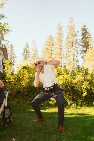 Someone wearing a hat and western-style outfit strikes a playful dance pose on a lawn with tall pine trees in background.