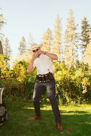 Person in western attire takes a dramatic golf swing on a sunny lawn with tall pine trees in the background.
