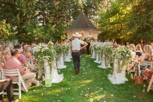 Outdoor garden wedding ceremony with floral aisle decor and white chairs under trees during golden hour.