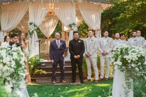 Outdoor wedding ceremony setup with white fabric drapes, floral arrangements, and string lights creating a romantic atmosphere.