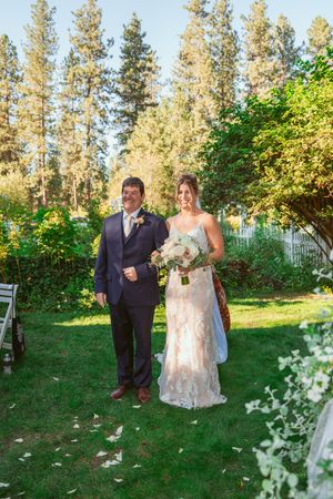 Outdoor wedding procession walking together on a green lawn path surrounded by tall evergreen trees on a sunny day.