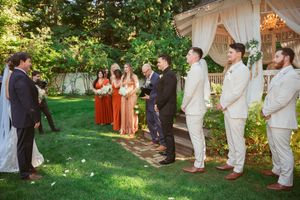 Wedding party gathers in a lush garden setting for outdoor ceremony photos under trees and natural sunlight.