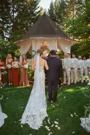 Outdoor garden wedding ceremony under rustic gazebo with white drapes and floral decorations in forest setting.