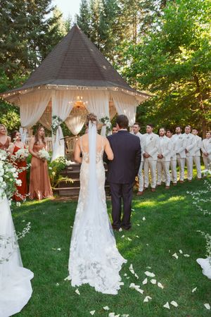 Outdoor wedding couple walking towards gazebo decorated with white drapes and flowers as wedding party looks on.