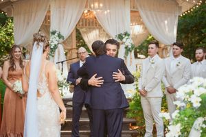 Two people embrace during an outdoor wedding ceremony under a white draped gazebo with wedding party looking on.