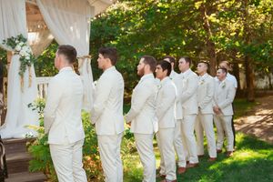 A wedding party in white attire stands in a line outdoors beneath trees and sunlight.