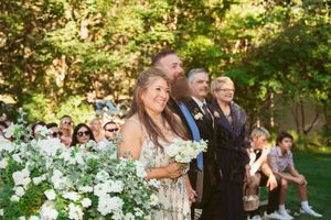 People gather outdoors at a wedding ceremony surrounded by white floral arrangements in a garden setting.