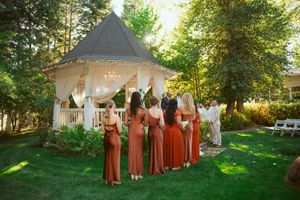 A wedding party in matching rust-colored dresses stands in an outdoor garden setting during a golden afternoon.
