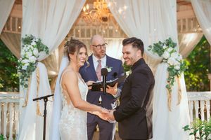 Happy couple stands at elegant outdoor wedding gazebo with white drapes and floral arrangements during ceremony.