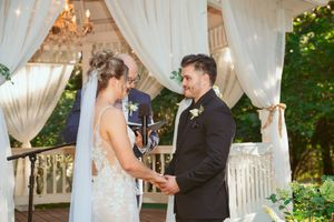 A couple exchanges vows under an elegant white draped gazebo decorated with twinkling lights and a chandelier.