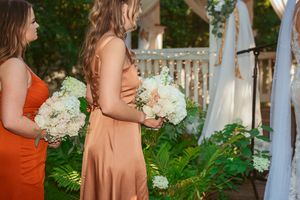 Bridesmaids in coral and blush dresses hold white floral bouquets while standing near greenery during outdoor wedding ceremony.