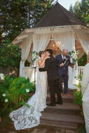 As the ceremony unfolds in an outdoor gazebo adorned with flowing white curtains and greenery, figures exchange vows.