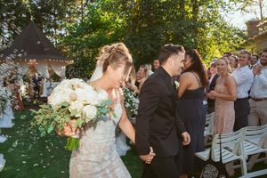 Newlyweds walk down outdoor aisle while guests throw flower petals during summer garden wedding ceremony.