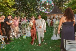 Couple walks down grassy outdoor aisle while guests blow bubbles during sunny garden wedding celebration.