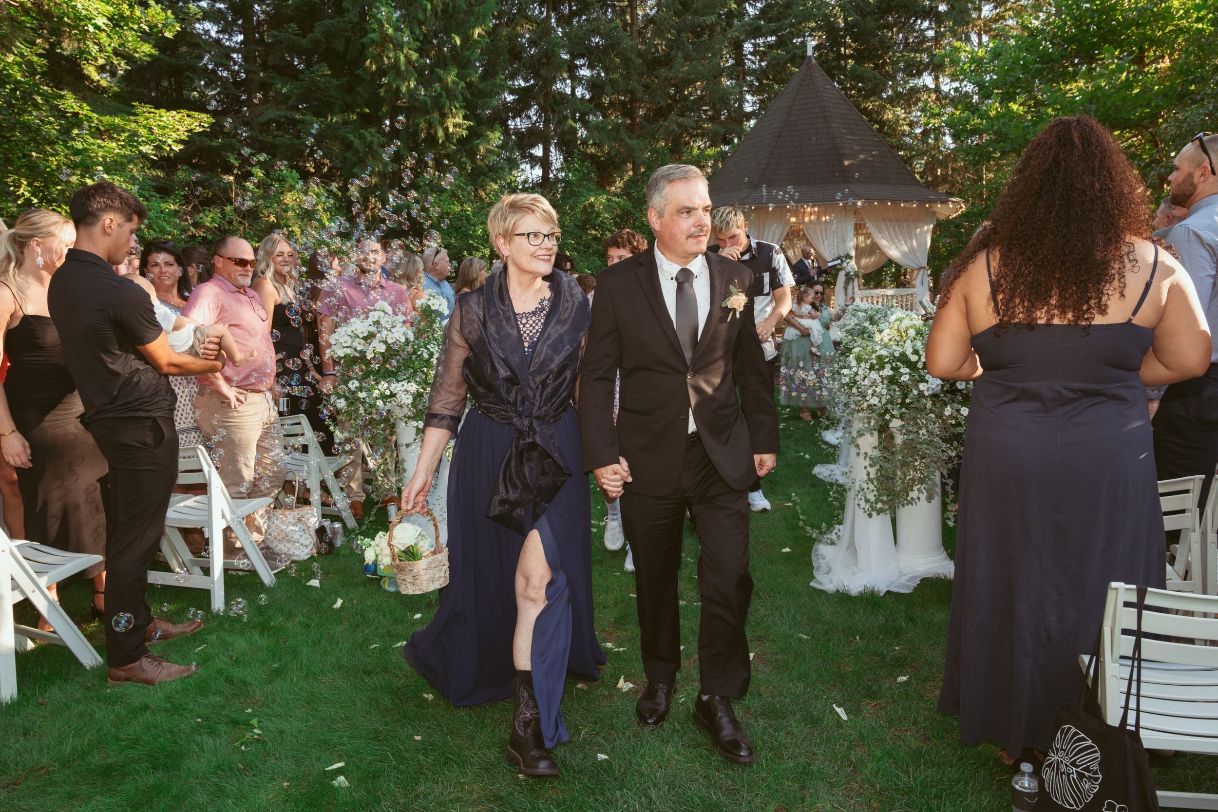 Wedding party members walk down the aisle at an outdoor garden ceremony with guests seated on white chairs.