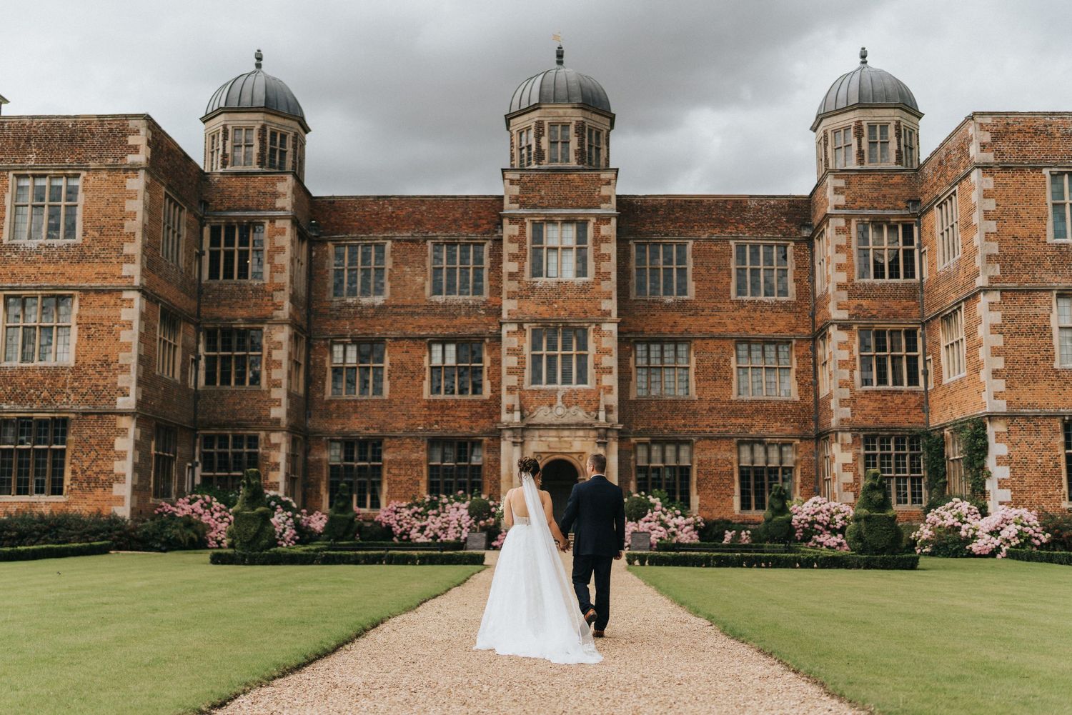 A wedding couple walking down a path toward a grand Tudor-style mansion with ornate domed towers and manicured lawn.