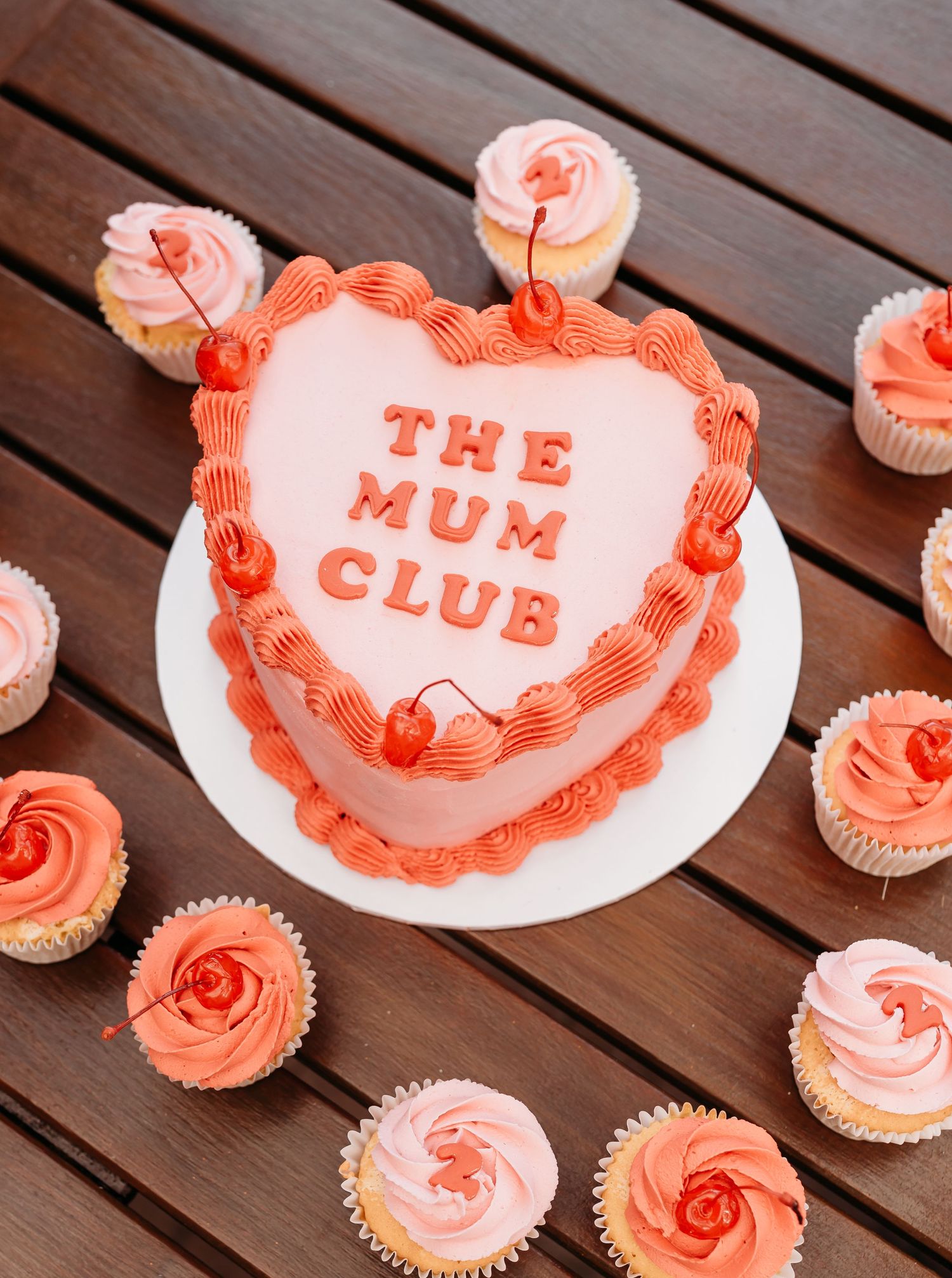 Heart-shaped pink cake decorated with 'The Mum Club' text and surrounded by matching coral-colored cupcakes on wooden deck.