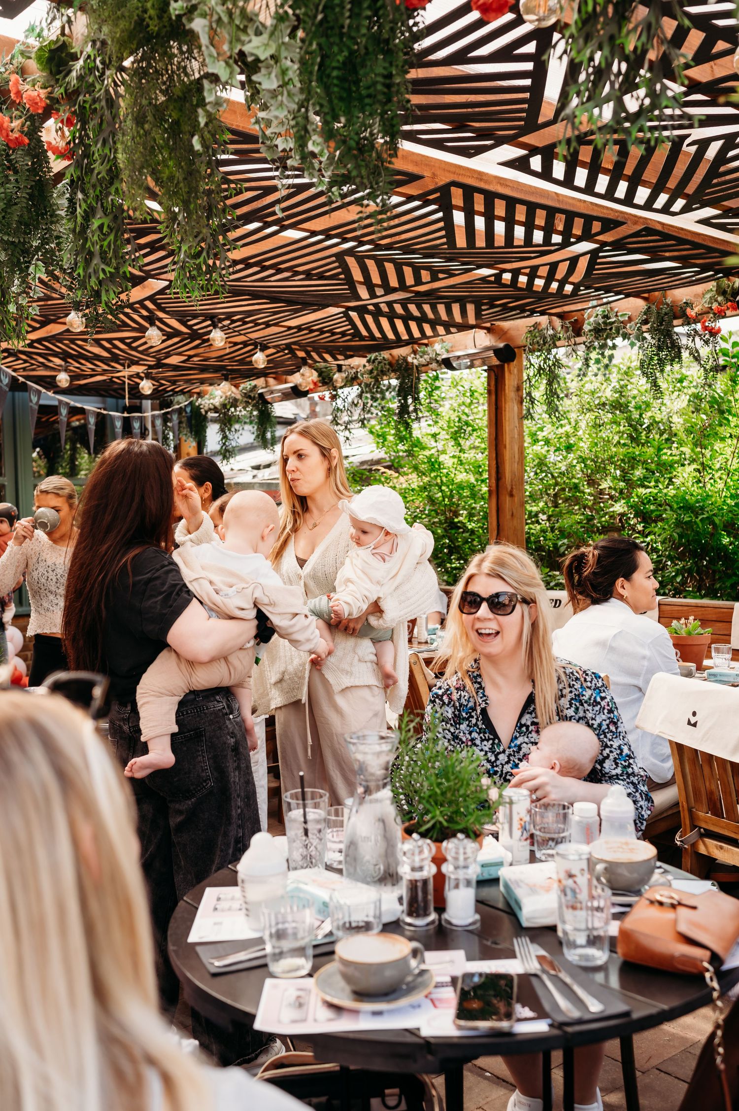 Outdoor restaurant patio with wooden pergola, hanging greenery and diners enjoying a social gathering at tables.