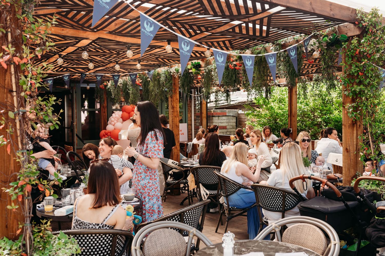 Outdoor restaurant patio with wooden pergola, string lights and patrons dining at tables surrounded by greenery.