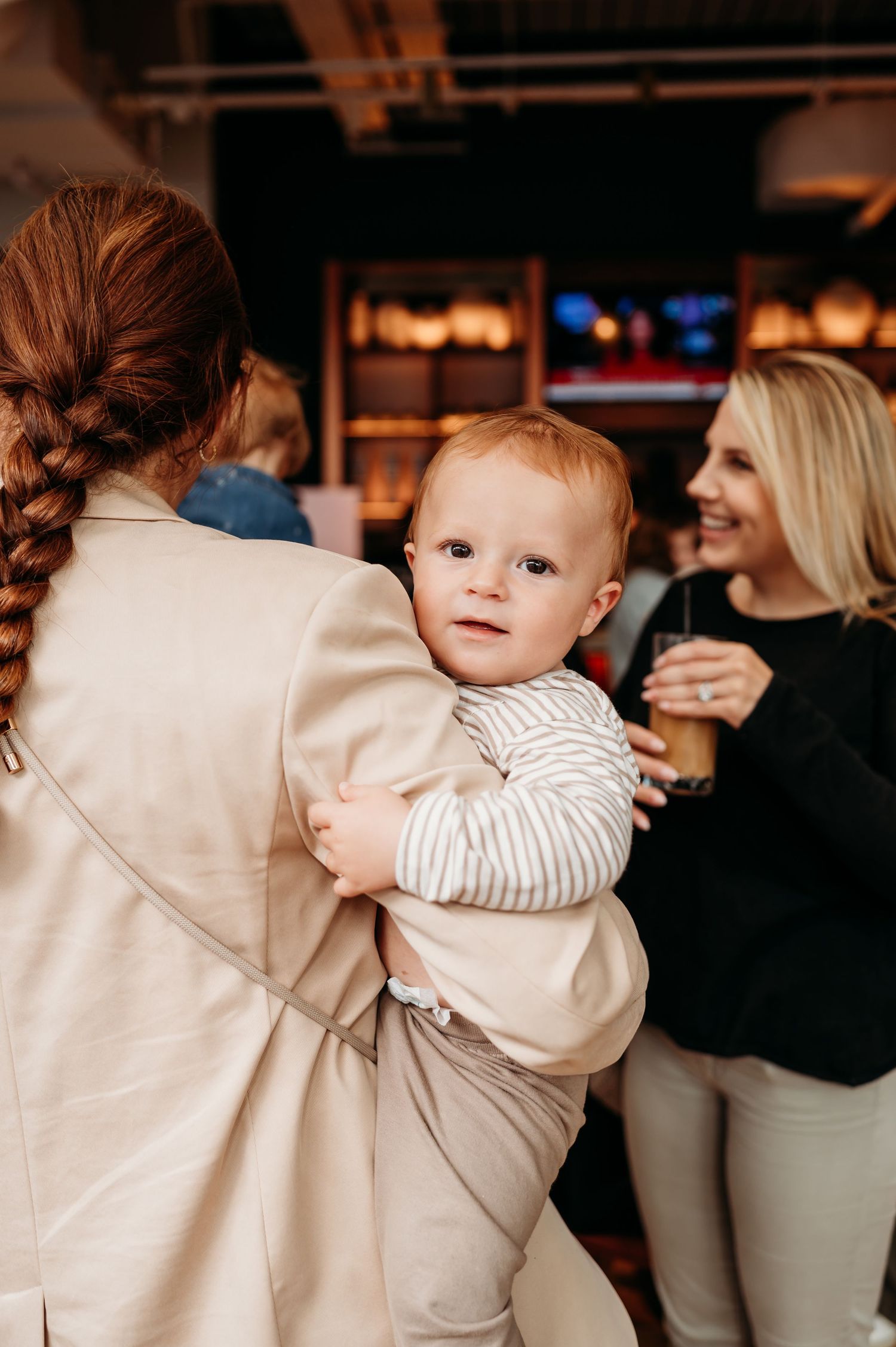 Baby in striped sweater reaches toward person wearing beige sweater in dimly lit indoor setting.