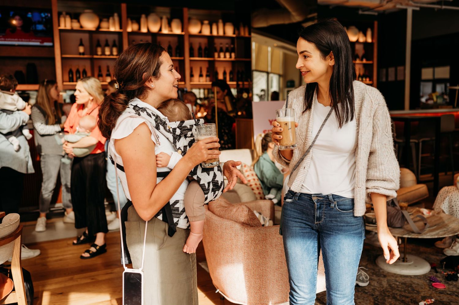 Two people enjoying drinks and conversation at a stylish bar while standing near wooden shelving with bottles.