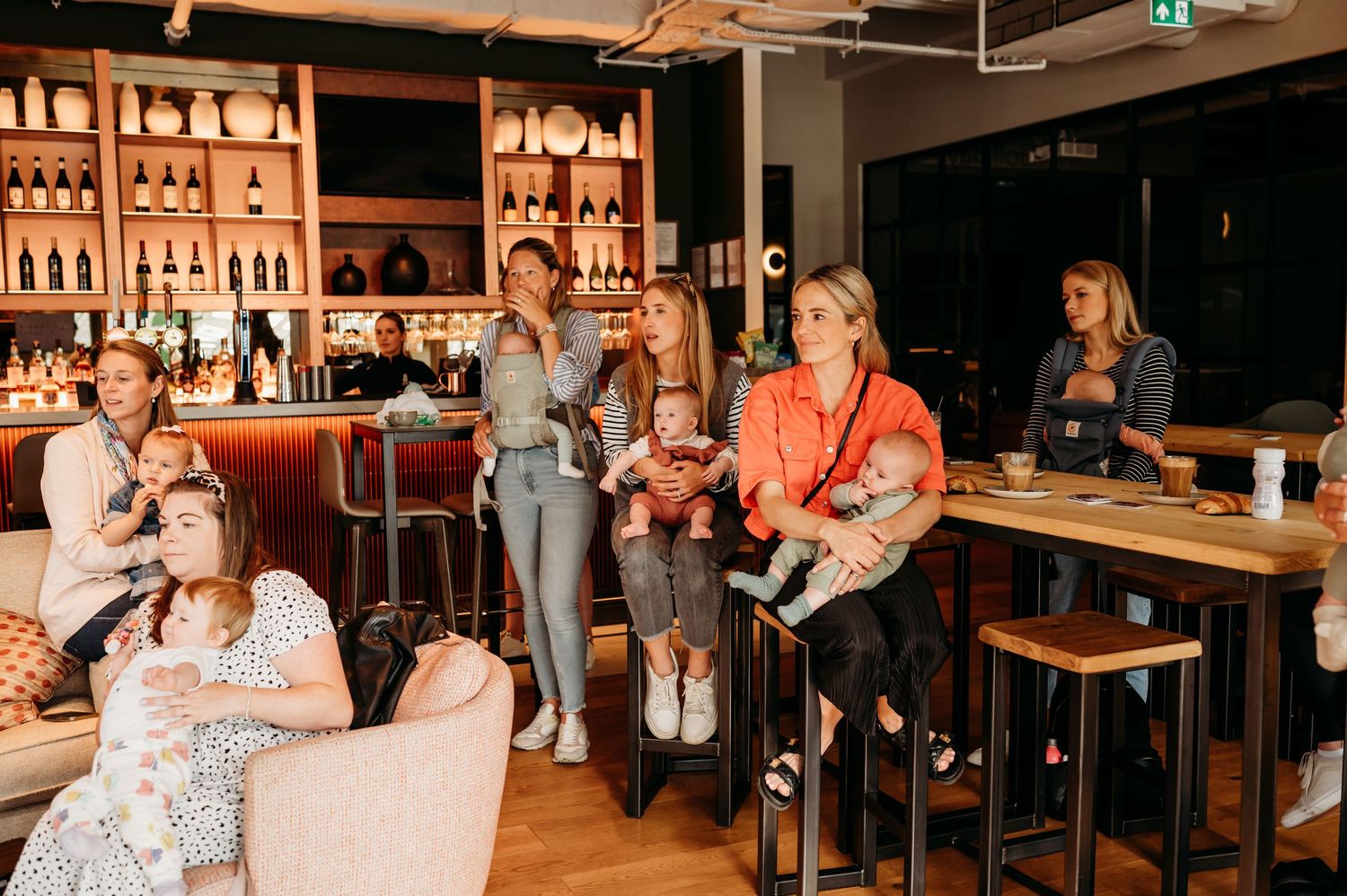 People gathered at a cozy bar with warm lighting, wooden stools, and shelves of bottles in the background.