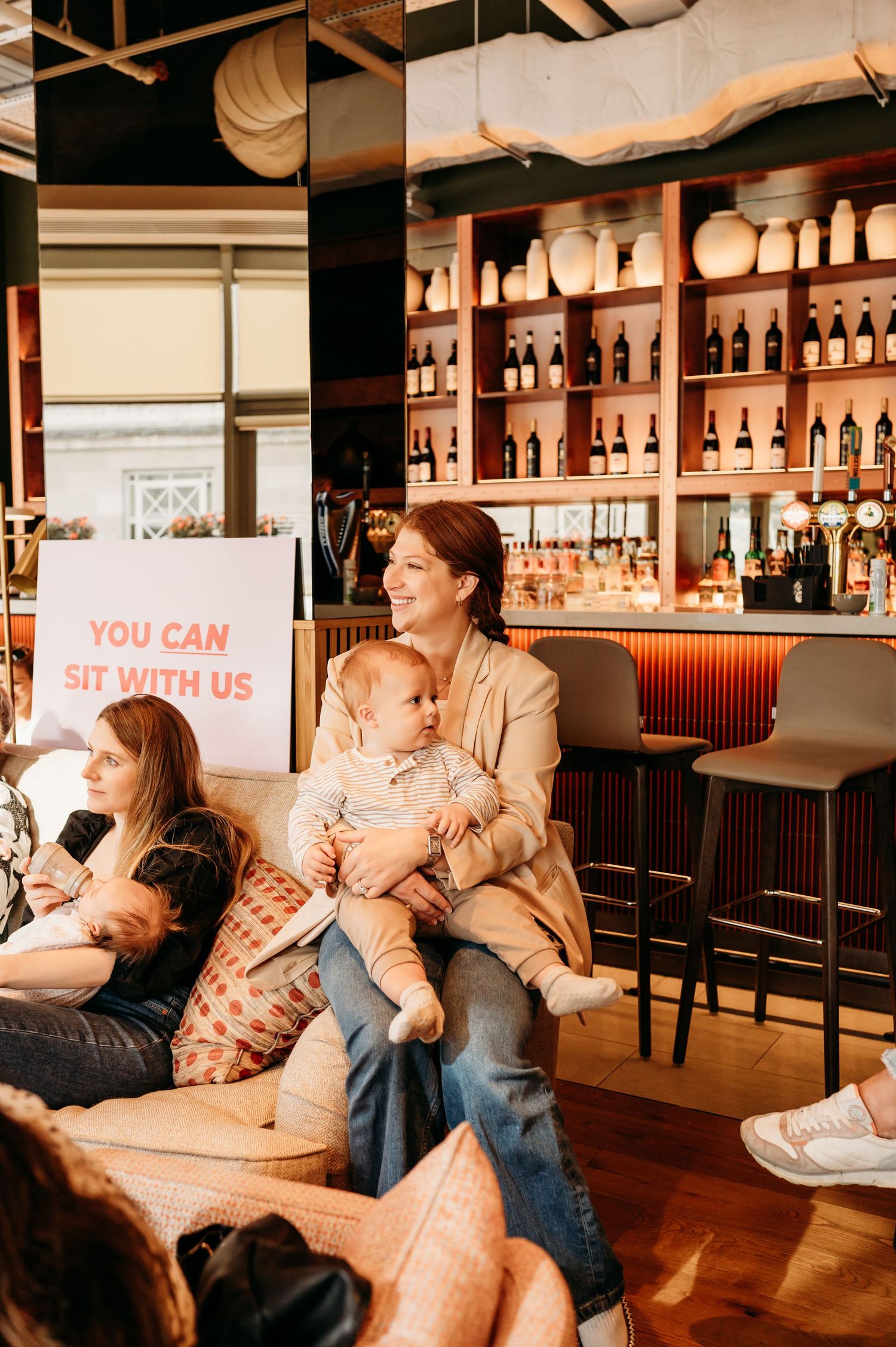 People gathered at a cozy bar interior with wooden decor, warm lighting and shelves of bottles behind the counter.