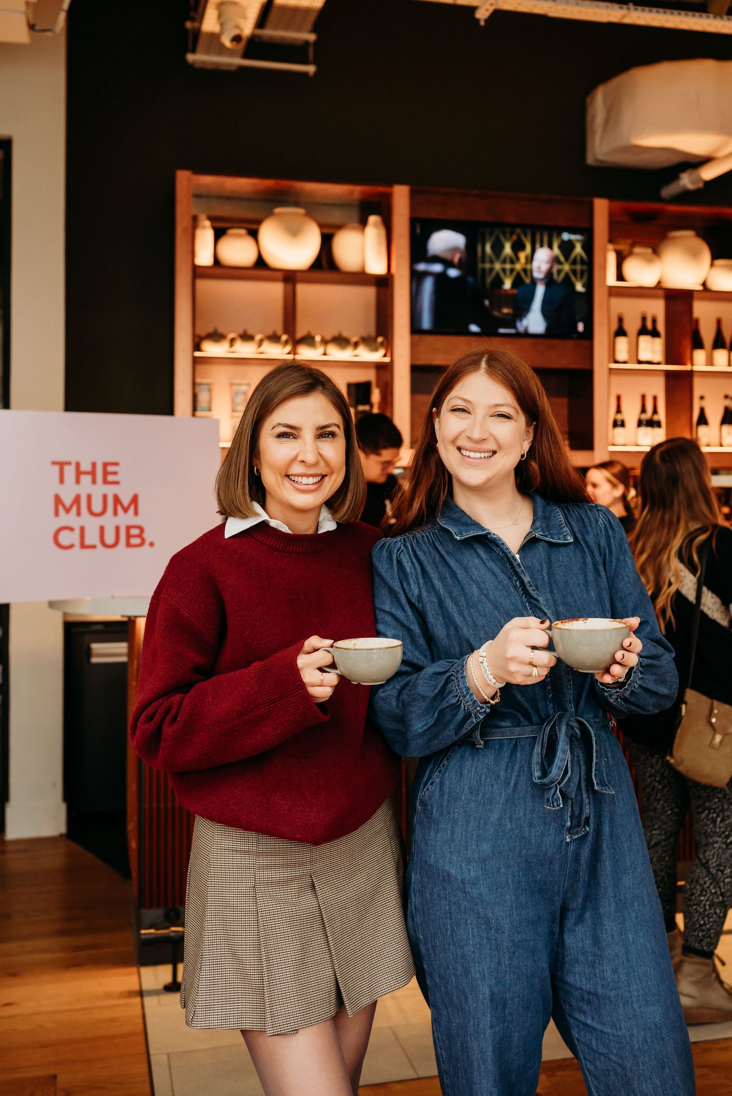 Two friends enjoy coffee together at The Mum Club, wearing a red sweater and denim jumpsuit.