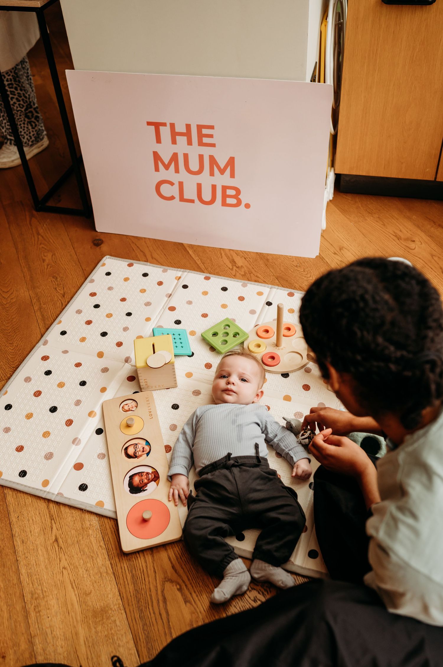 A baby lies on a polka dot play mat next to educational toys while someone adjusts their clothing.
