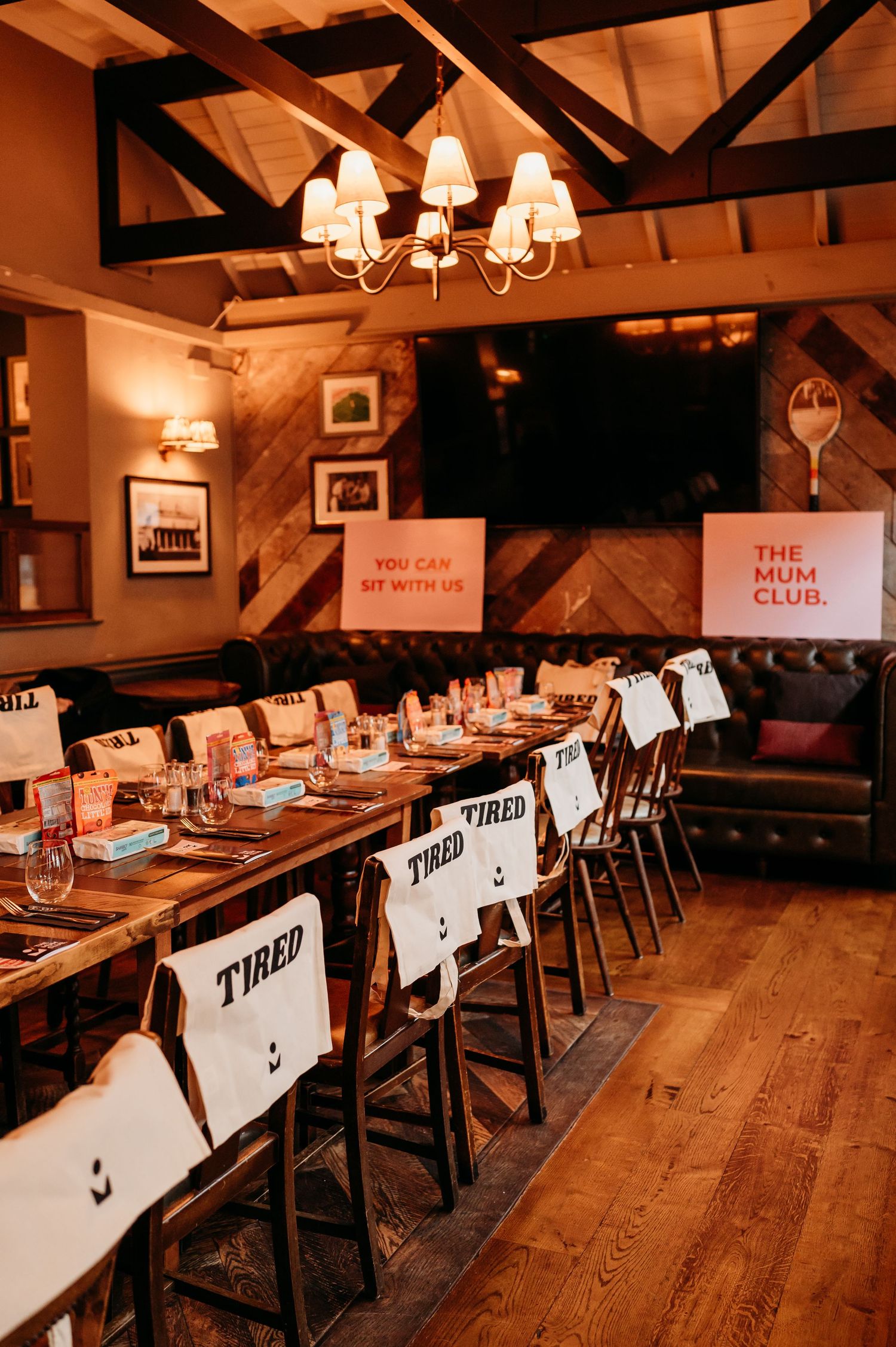 Rustic restaurant interior with wooden tables labeled 'TIRED' and warm lighting from chandelier fixtures.