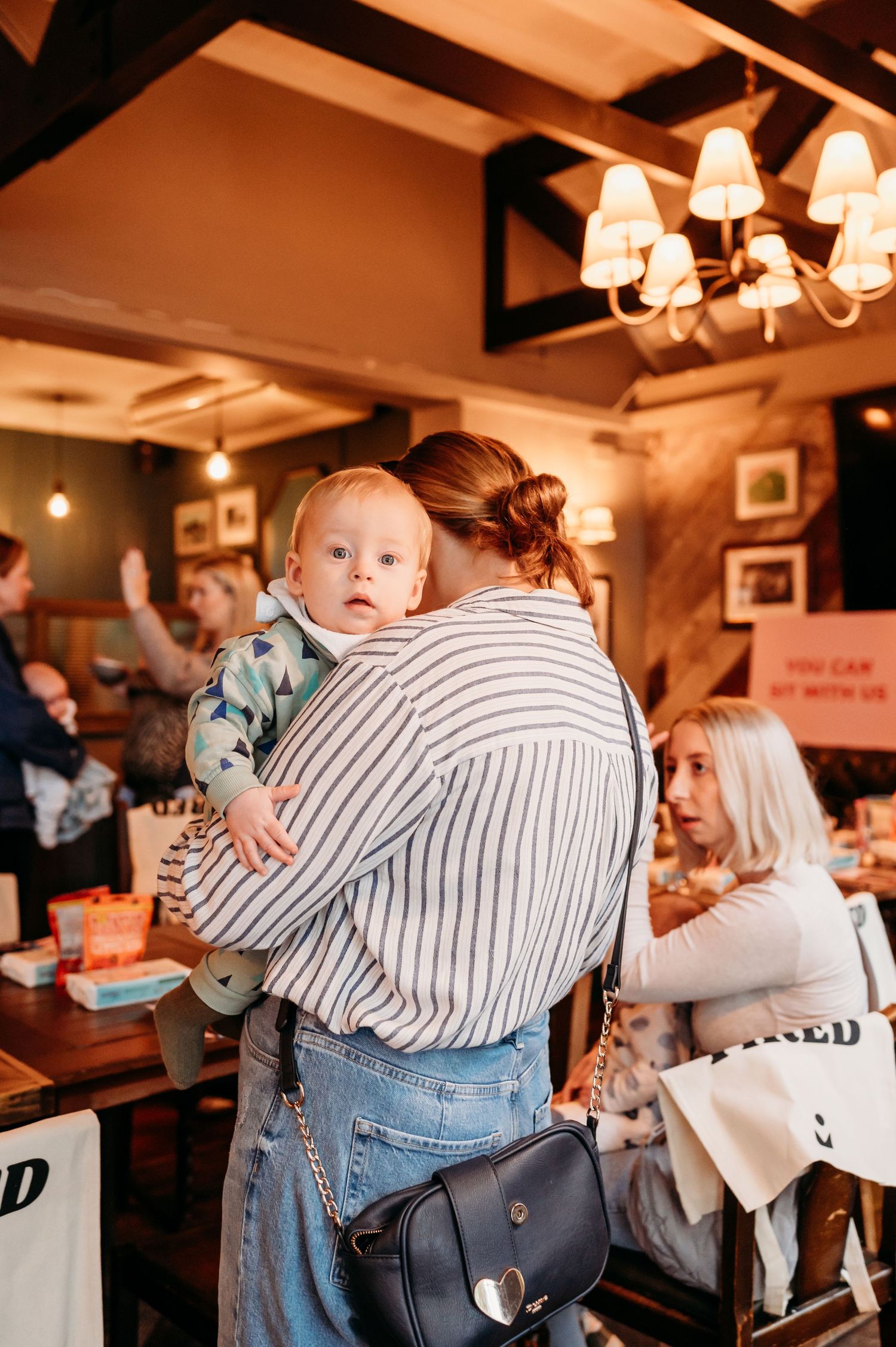 Cozy restaurant scene showing someone in a striped shirt holding a baby while others dine in warm lighting.