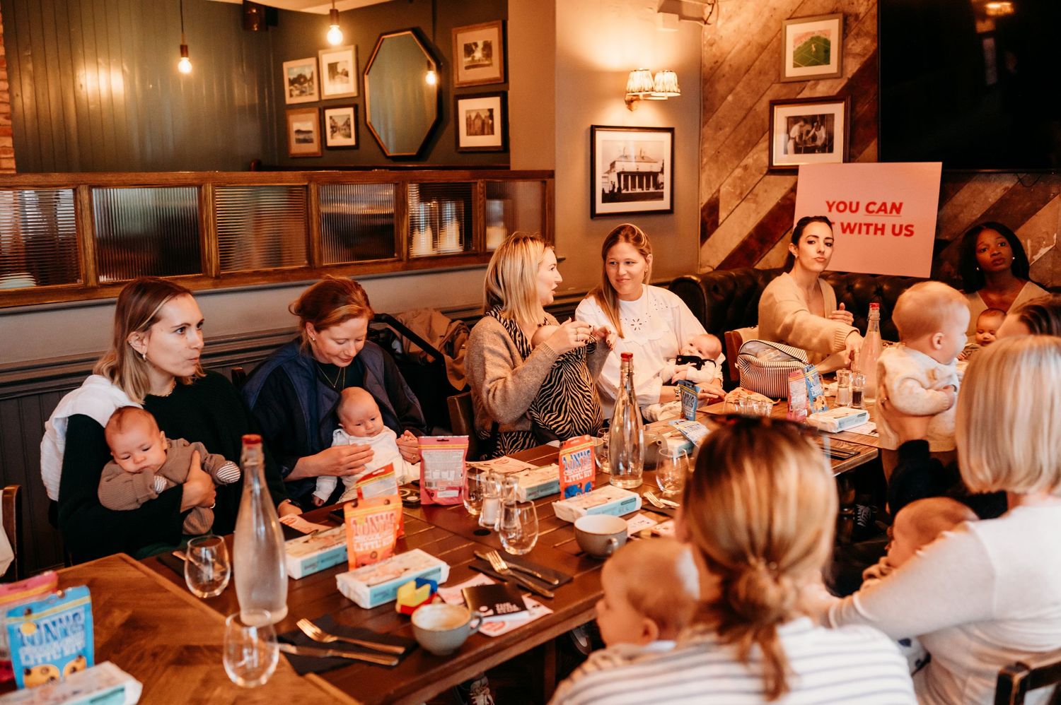 People gathered around a wooden table enjoying food and drinks in a cozy restaurant with vintage decor and warm lighting.