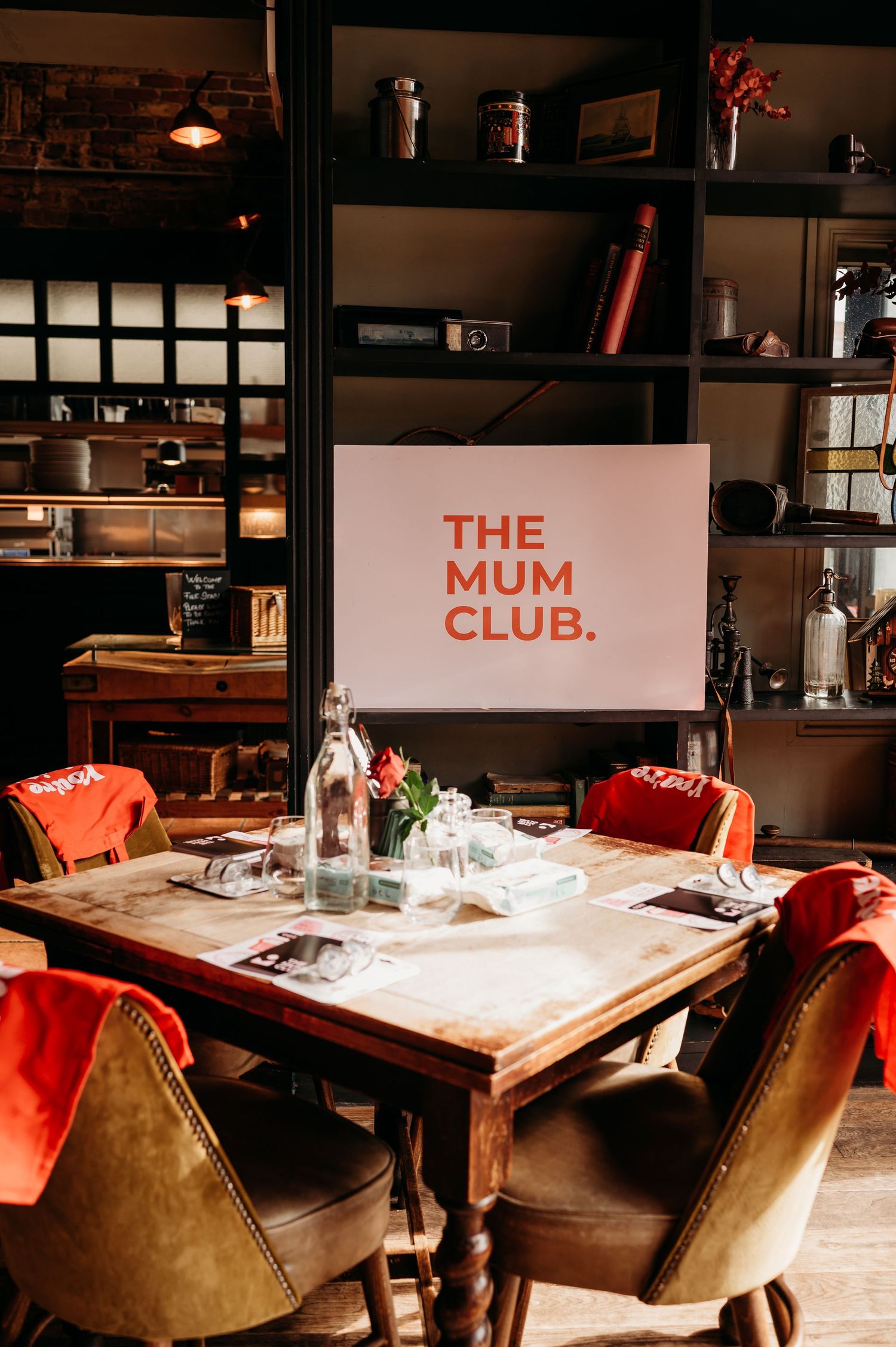 Cozy restaurant interior with a wooden table, red velvet chairs, and a sign reading 'THE MUM CLUB' on display.